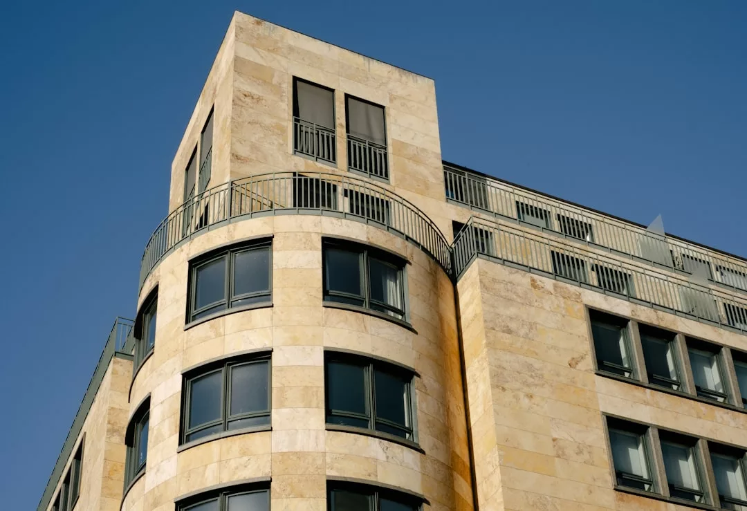 Das moderne mehrstöckige Gebäude mit seiner beigen Steinfassade, den abgerundeten Ecken, großen Fenstern und Metallgeländern erinnert an die zeitgenössische Eleganz Mallorcas vor dem Hintergrund eines klaren blauen Himmels.
