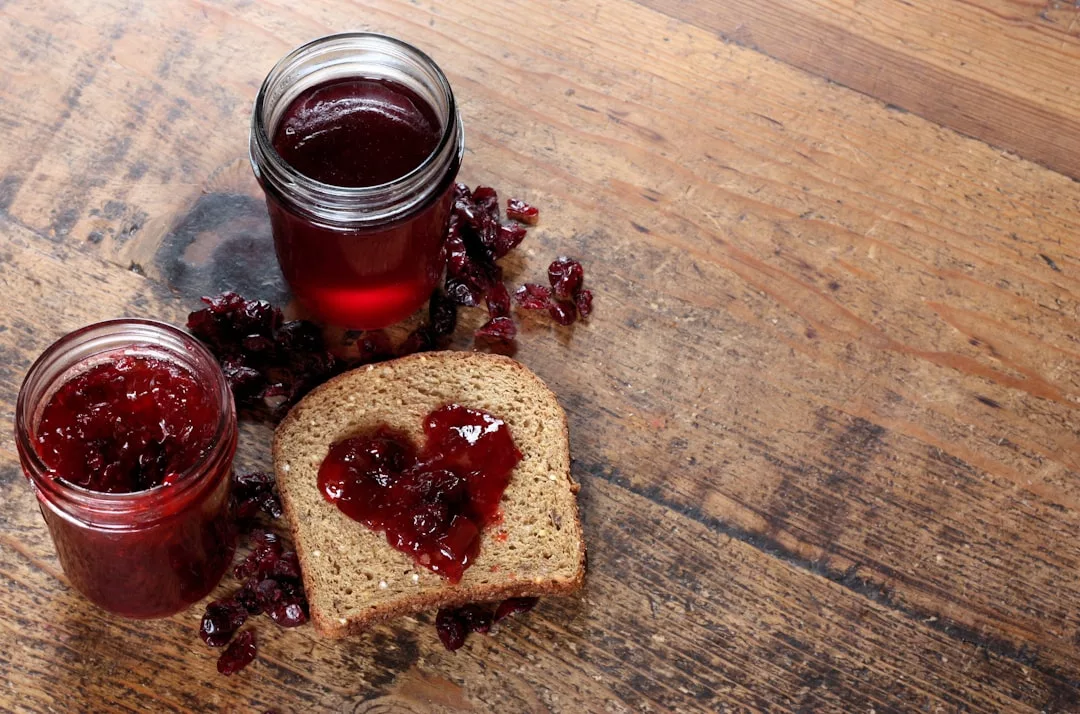 Zwei Gläser mit roter Marmelade und getrockneten Preiselbeeren stehen auf einem Holztisch, auf dem eine Scheibe Brot in Herzform mit Mallorca-Marmelade belegt ist.