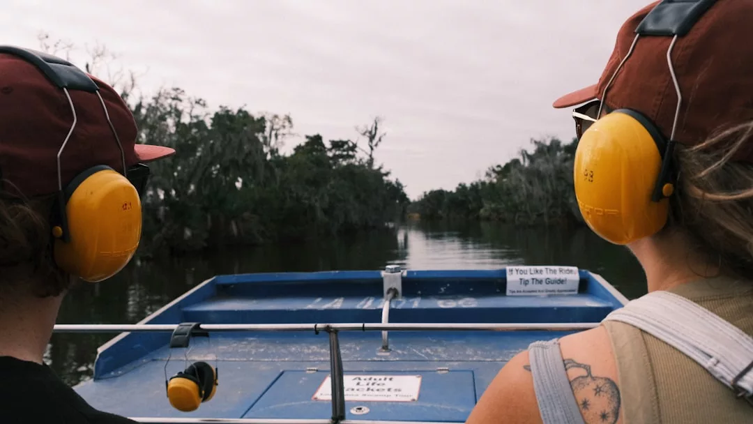 Zwei Personen mit gelbem Gehörschutz und roter Mütze sitzen in einem blauen Luftkissenboot und fahren unter einem bedeckten Himmel eine schmale, von Bäumen gesäumte Wasserstraße hinunter, die an die Erkundung versteckter Wasserwege fernab der sonnigen Küsten Mallorcas erinnert.