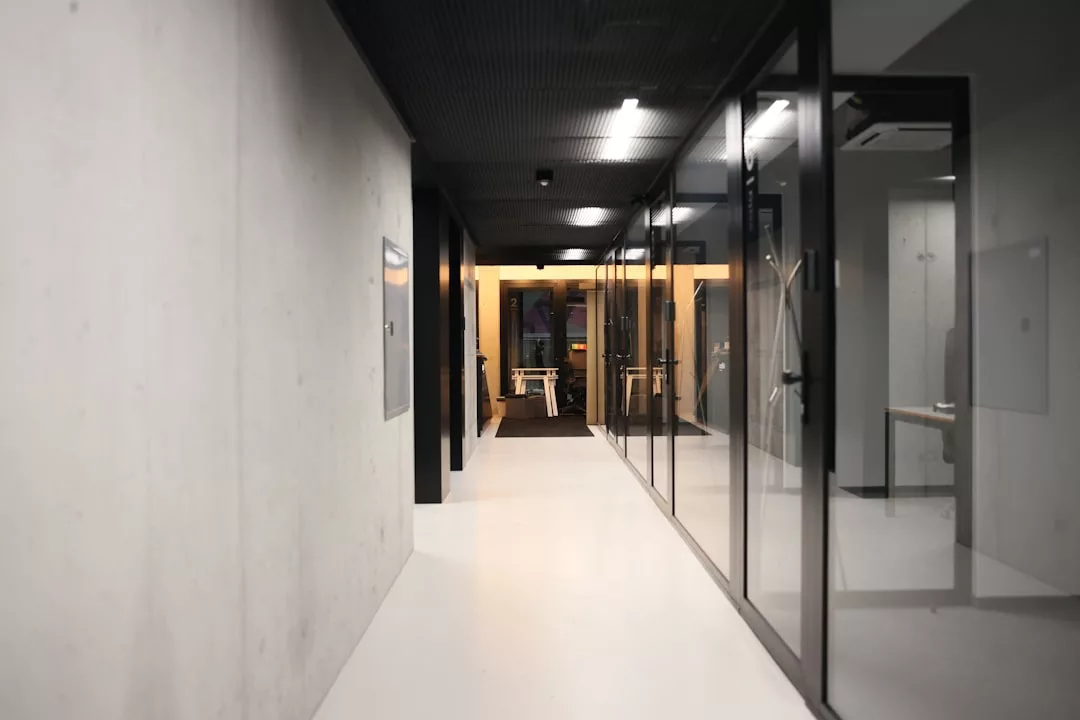 Moderner Büroflur mit weißem Boden, gläsernen Besprechungsräumen auf der rechten Seite und einer Betonwand auf der linken Seite, die zu einem Bereich mit Schreibtischen und warmer, von Mallorca inspirierter Beleuchtung führt.