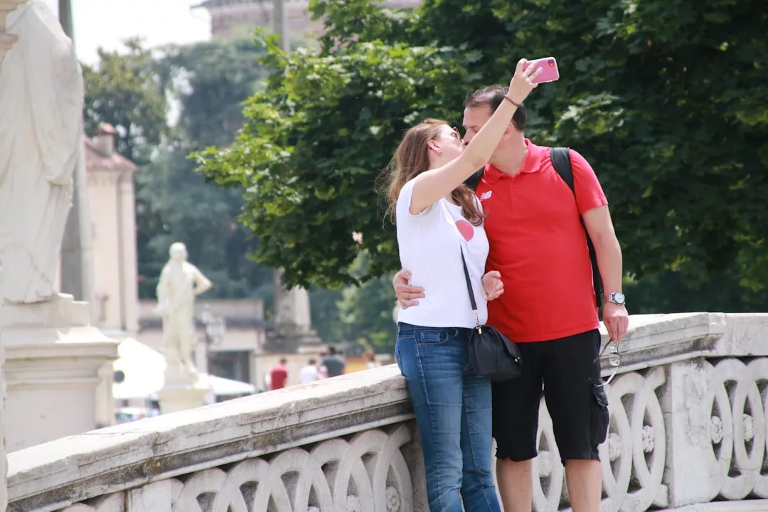 Eine Frau und ein Mann stehen dicht beieinander auf einer Steinbrücke auf Mallorca und machen ein Selfie mit einem Smartphone; im Hintergrund sind Statuen und Bäume zu sehen.