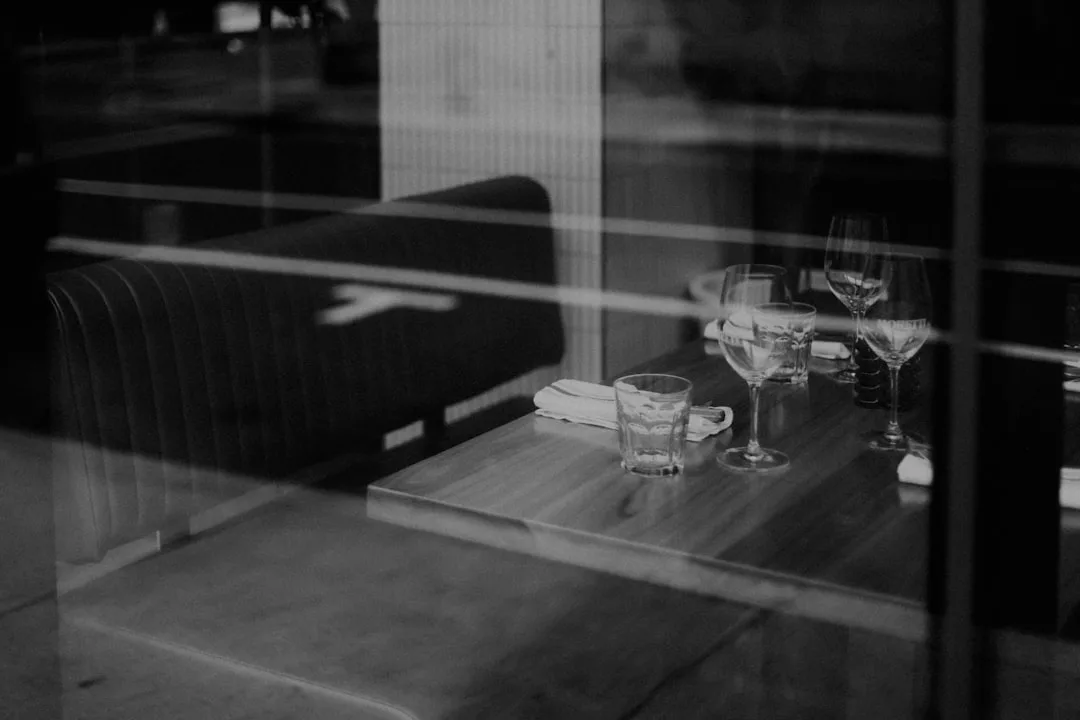 Schwarz-Weiß-Foto eines Restaurantstandes auf Mallorca, gesehen durch ein Fenster, mit einem gedeckten Tisch mit Gläsern, Servietten und Besteck auf poliertem Holz.