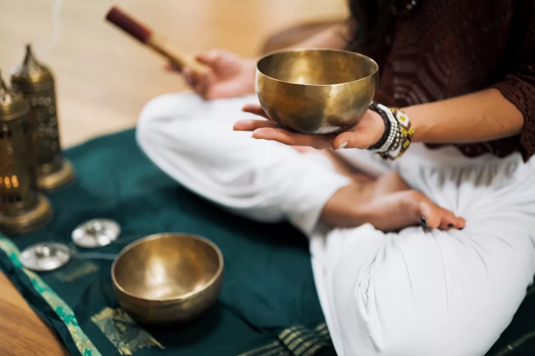 Im Schneidersitz auf einer Matte sitzende Person auf Mallorca, die eine Klangschale mit einer weiteren Schale, einem Schlegel und Laternen in der Nähe hält, was auf eine Meditations- oder Klangheilungspraxis hindeutet.