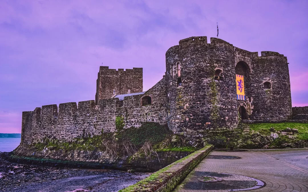 Eine große Steinburg mit hohen Mauern und einem runden Turm steht in der Nähe des Wassers unter einem violetten Himmel auf Mallorca. Ein buntes Banner hängt an der Hauptwand über dem Eingang.