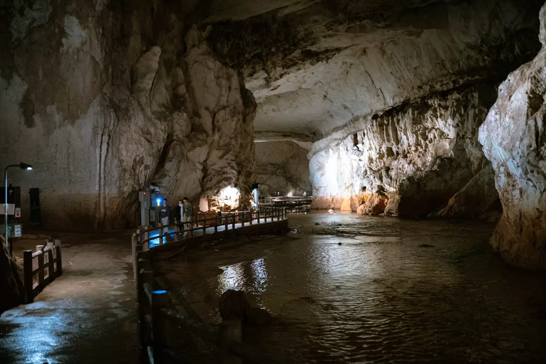 Ein Holzsteg führt durch eine große, beleuchtete Höhle mit felsigen Wänden und einem flachen Bach, der unter ihr hindurchfließt, auf Mallorca.