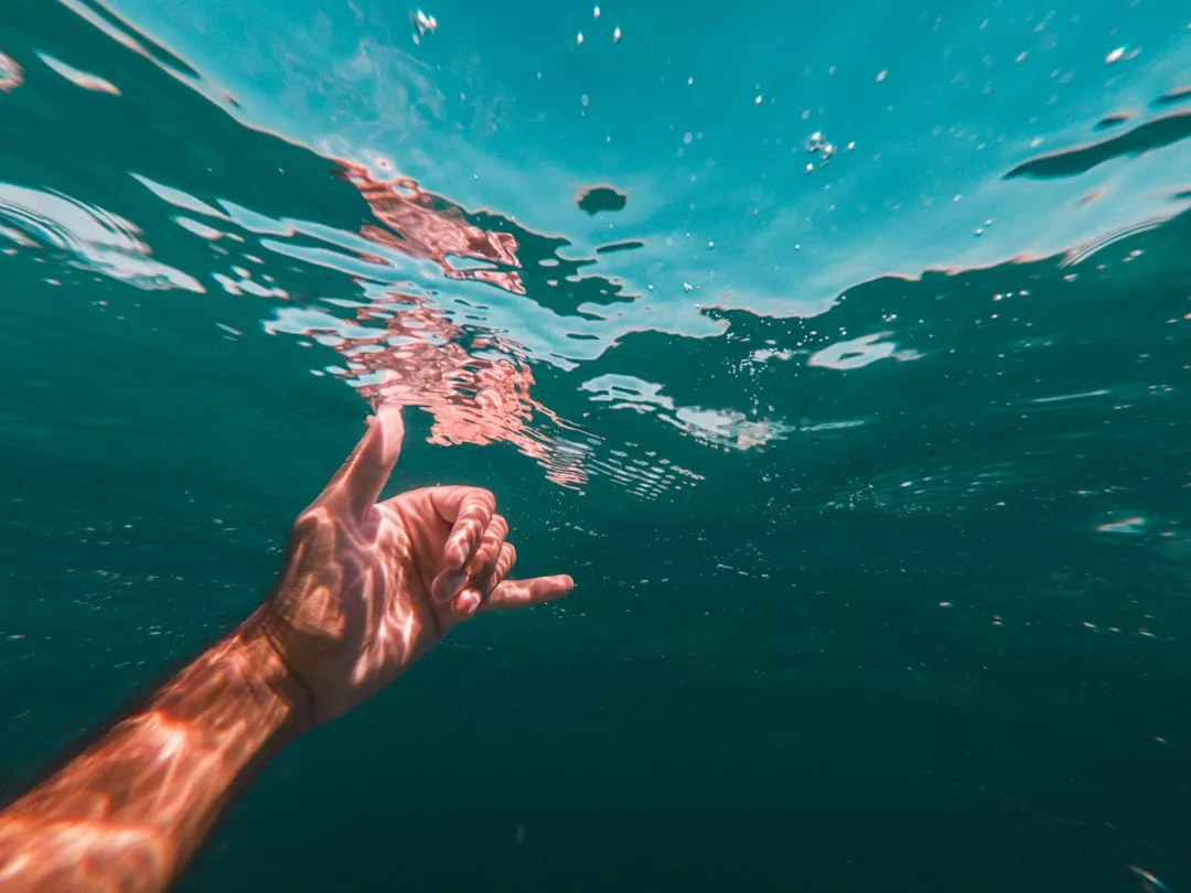 Eine Hand, die das Shaka-Zeichen macht, ist vor der Küste Mallorcas teilweise unter Wasser getaucht, wobei sich das Sonnenlicht auf der Haut und der glitzernden Wasseroberfläche spiegelt.