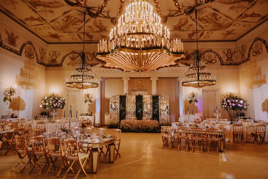 Eleganter Bankettsaal mit verzierten Kronleuchtern, dekorierter Decke, runden und rechteckigen Tischen für eine Veranstaltung und einer Blumenkulisse auf der Bühne - perfekt für eine von Mallorca inspirierte Feier.