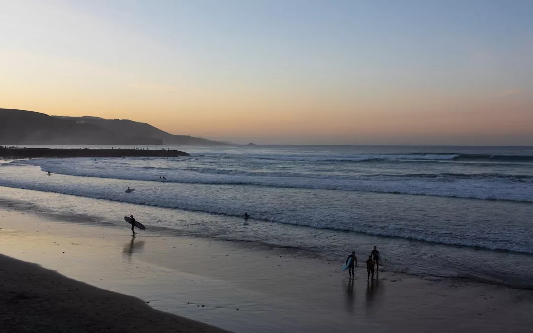 Surfer und Strandbesucher genießen den Sandstrand Mallorcas mit sanften Wellen bei Sonnenuntergang; im Hintergrund sind Hügel und ein Wellenbrecher zu sehen.