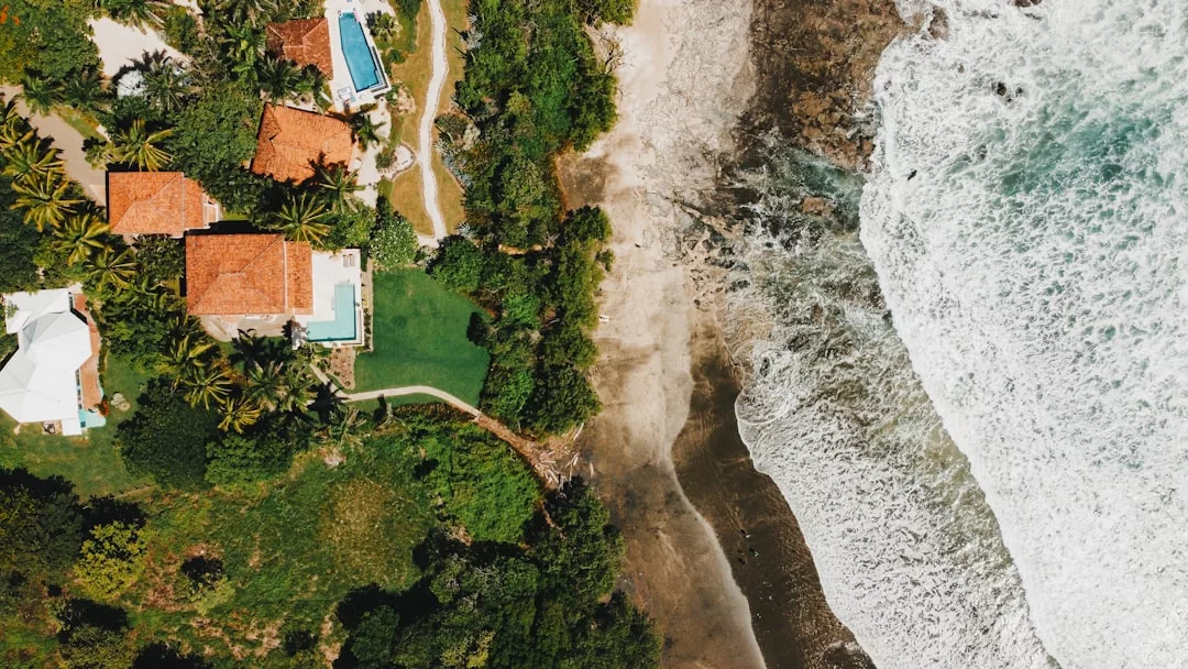 Luftaufnahme von Häusern mit roten Dächern und Swimmingpools inmitten von grünen Bäumen auf Mallorca, angrenzend an einen Sandstrand und die Wellen des Meeres.