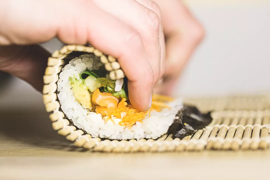 Nahaufnahme von Händen, die eine Bambusmatte benutzen, um Sushi mit Reis, Gemüse und Seetang auf einer hölzernen Oberfläche zu rollen, inspiriert von den frischen Aromen Mallorcas.