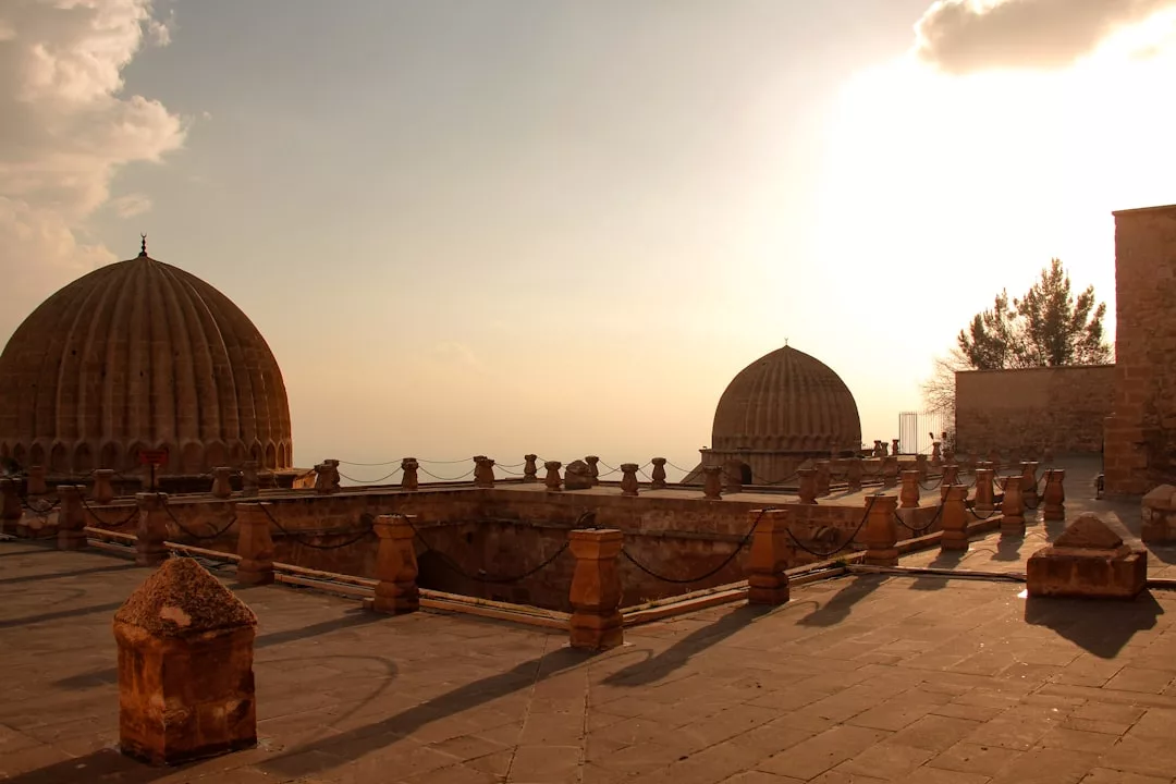 Die Sonne geht über einem alten Steinhof auf Mallorca unter, in dem zwei Kuppelbauten stehen, die von Säulen und Ketten begrenzt werden und lange Schatten auf den Boden werfen.