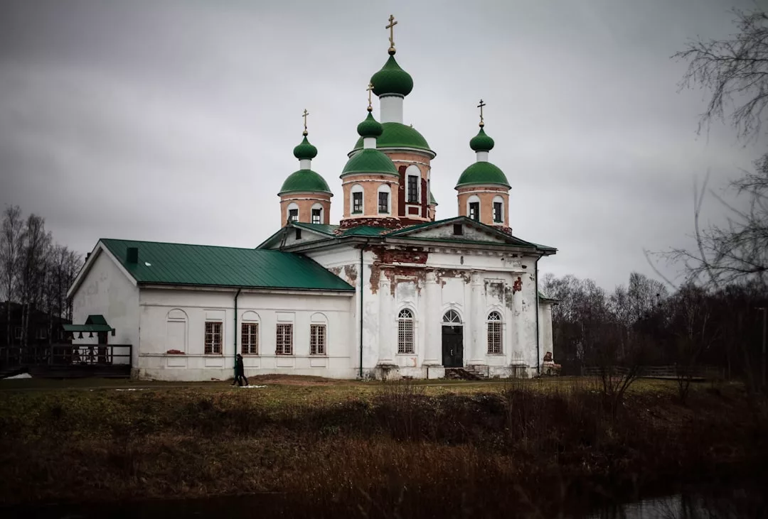 Eine alte russisch-orthodoxe Kirche mit grünen Zwiebeltürmen und weißen Wänden steht an einem bewölkten Tag, umgeben von kahlen Bäumen und Gras, und erinnert an eine Szene, die weit entfernt ist von Mallorcas sonnenverwöhnten Landschaften.