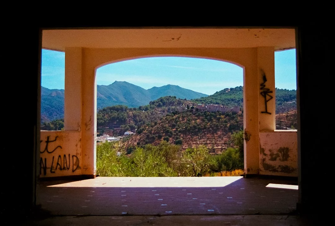 Durch eine große rechteckige Öffnung in einem mit Graffiti beschmierten, verlassenen Gebäude eröffnet sich ein Blick auf die grünen Hügel Mallorcas und eine kleine Stadt.