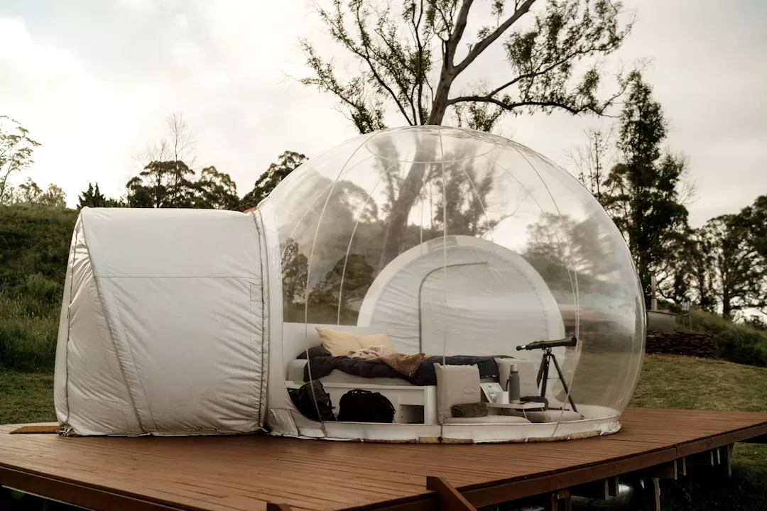 Ein transparentes Blasenzelt steht auf einer Holzplattform im Freien auf Mallorca, ausgestattet mit einem Bett, Kissen und einem Teleskop, umgeben von Bäumen und Gras.