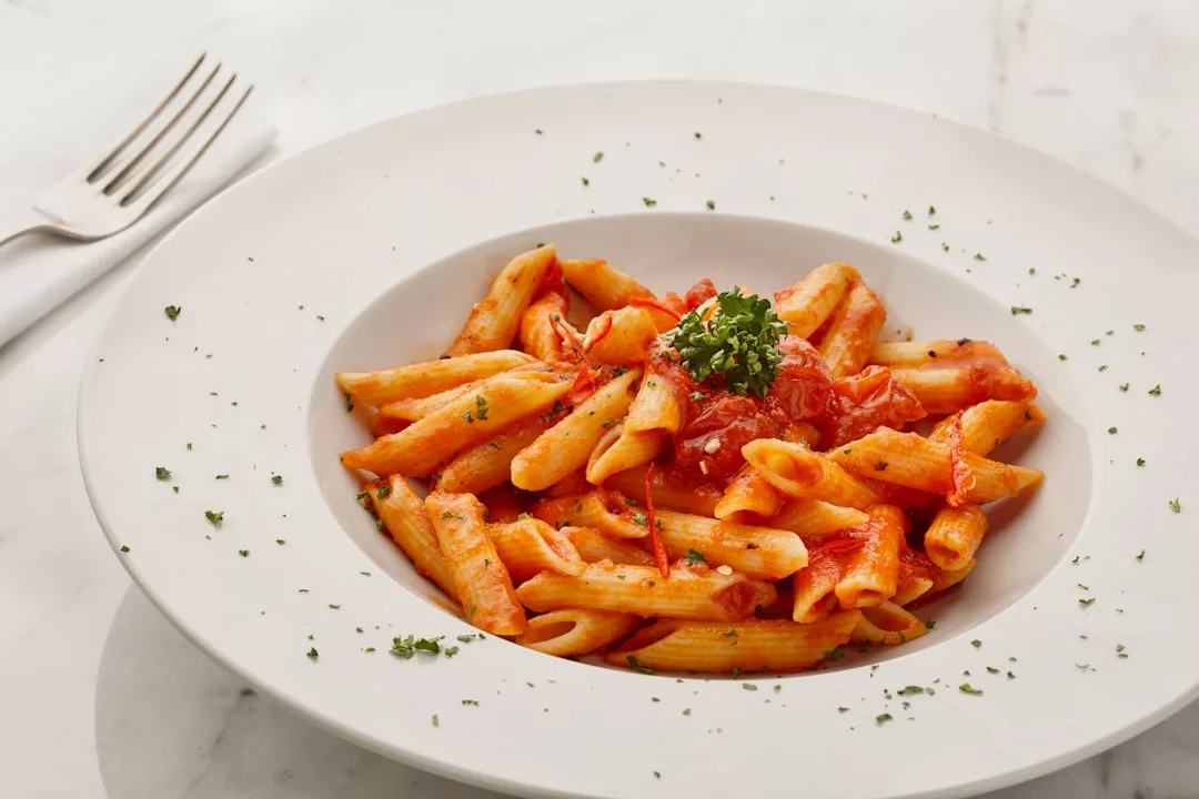 Ein weißer Teller mit Penne in Tomatensauce, garniert mit gehackten Kräutern und einem Zweig Petersilie, steht auf einer Marmorplatte neben einer Gabel und einer Serviette und bringt einen Hauch von Mallorca an Ihren Tisch.