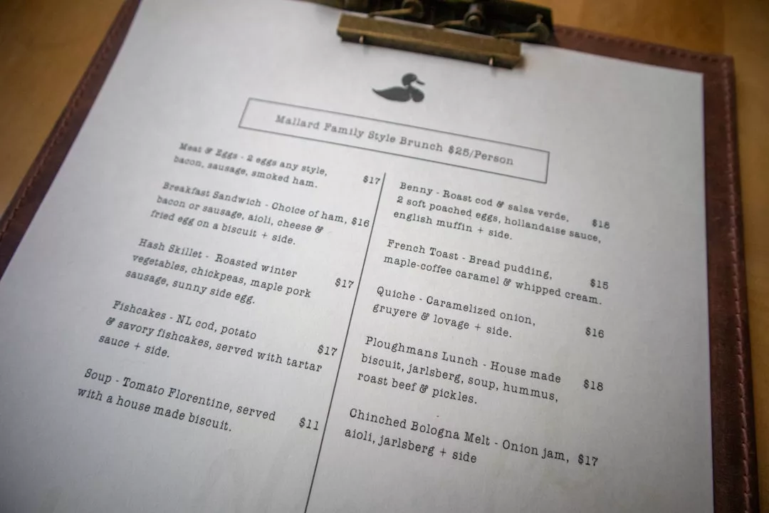 Nahaufnahme eines Brunch-Menüs auf einem Klemmbrett mit verschiedenen Gerichten und Preisen, darunter Mallorca-Sandwiches, Eier, Haschee, Suppe, Fischfrikadellen und Desserts.
