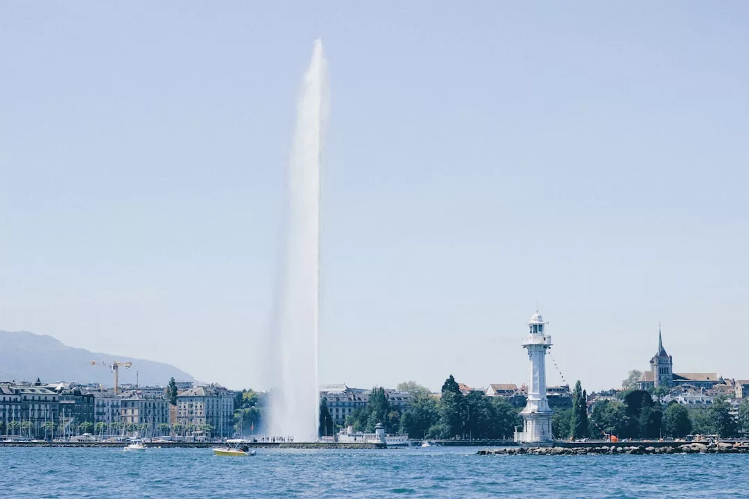 Ein großer Springbrunnenstrahl fließt neben einem weißen Leuchtturm an der Küste Mallorcas in die Höhe. Im Hintergrund sind die Gebäude der Stadt und die fernen Berge unter einem klaren Himmel zu sehen.
