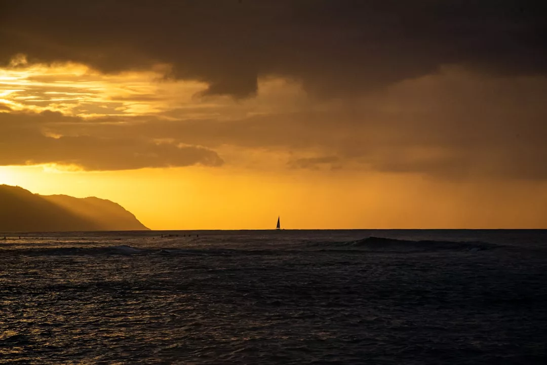 Ein Segelboot ist am Horizont unter einem dramatischen goldenen Sonnenuntergangshimmel vor der Küste Mallorcas zu sehen, mit dunklen Wolken darüber und ruhigen Meereswellen im Vordergrund.