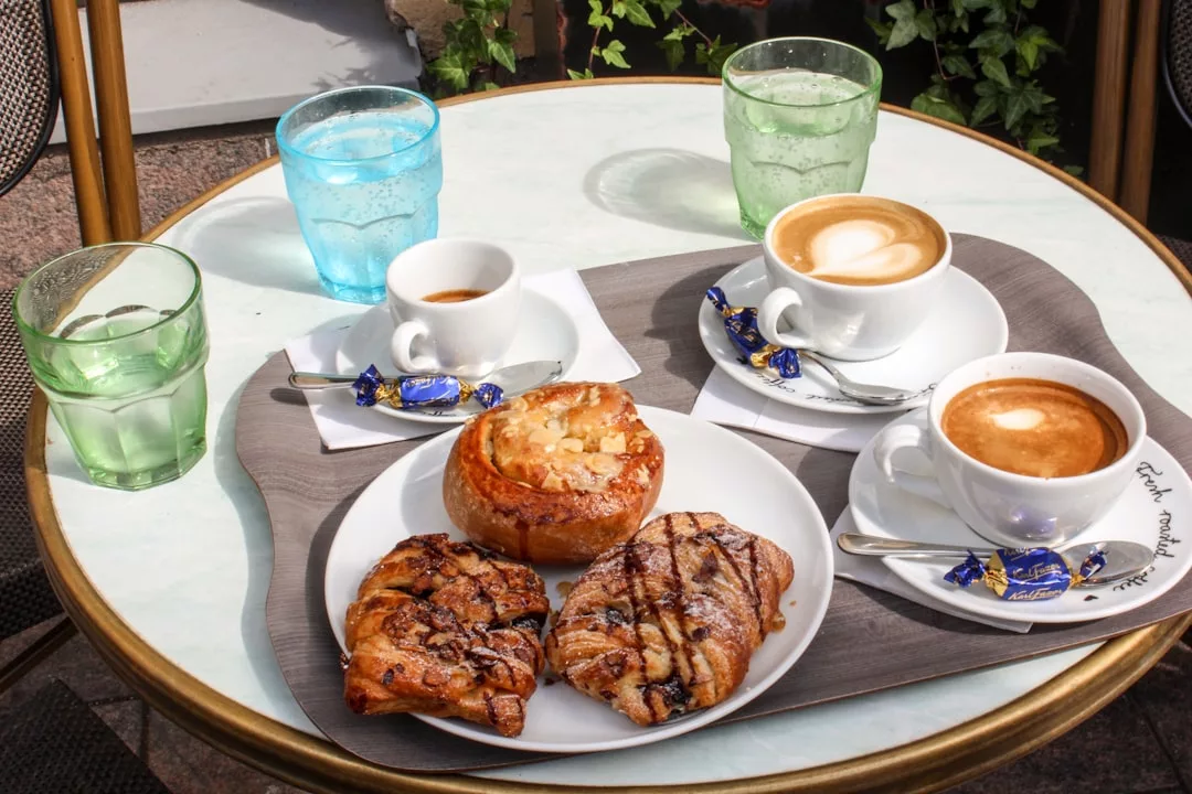 Ein runder Tisch auf Mallorca mit zwei Cappuccinos, einem Espresso, einem Gebäck, zwei Croissants, zwei Gläsern Wasser und verpackten Pralinen auf einem Tablett im Freien.