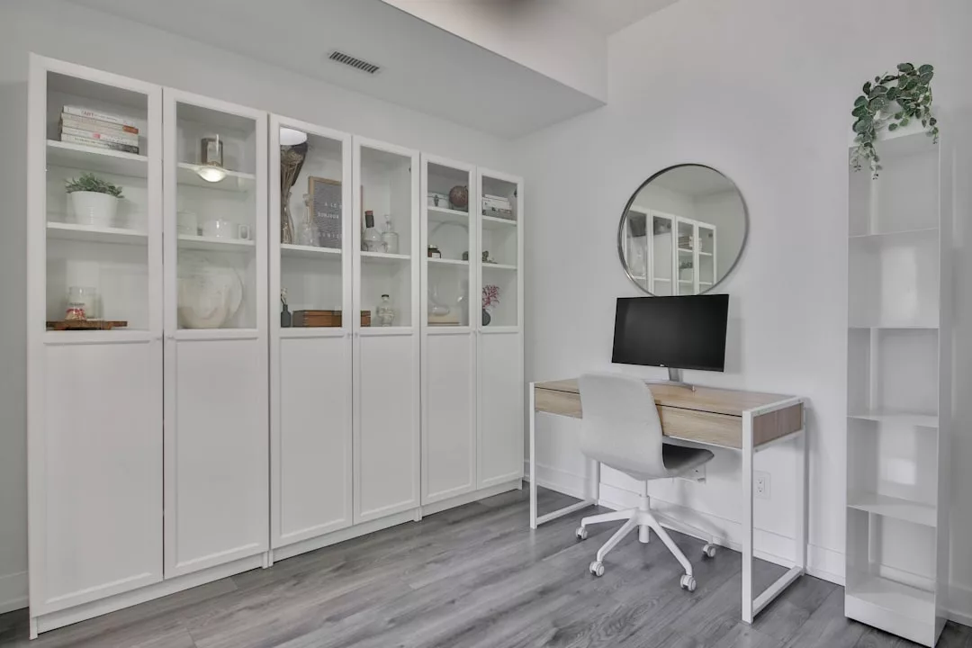 Modernes Heimbüro mit weißem Schreibtisch, Computermonitor, Drehstuhl und rundem Wandspiegel. Weiße Regale mit Glastüren präsentieren Bücher, Pflanzen und Dekoration und bringen einen Hauch von mallorquinischer Ruhe in den Raum.
