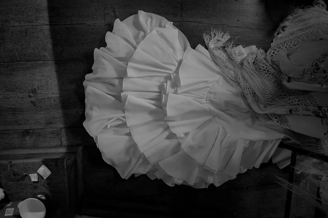 Ein weißes, gerüschtes Kleid mit mehrlagigem Stoff liegt auf dem Holzboden und erinnert an den sonnenverwöhnten Charme Mallorcas, daneben liegen ein fransenbesetzter Schal und verstreute kleine Gegenstände.
