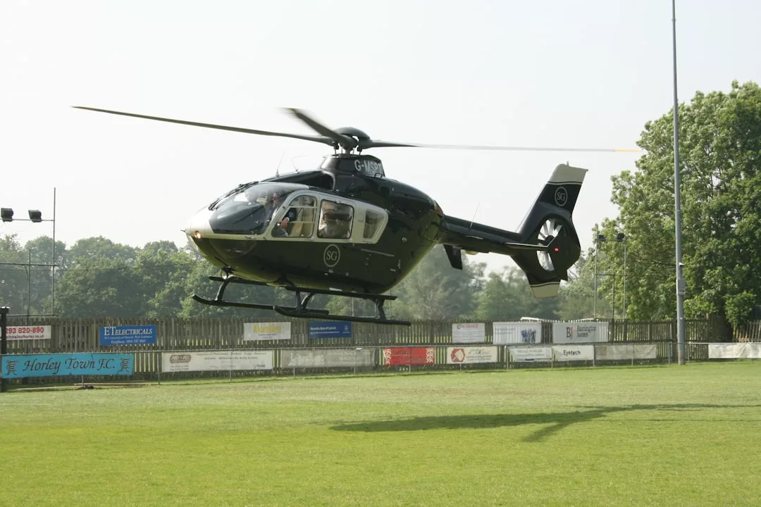 Ein grün-weißer Hubschrauber schwebt über einer Wiese auf Mallorca, im Hintergrund sind Bäume und Werbetafeln zu sehen.