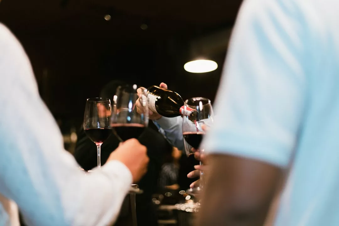 Bei einem lebhaften Treffen auf Mallorca schenkt eine Person Rotwein in Gläser ein, die von anderen gehalten werden, während im Vordergrund Personen in weißen Hemden zu sehen sind.