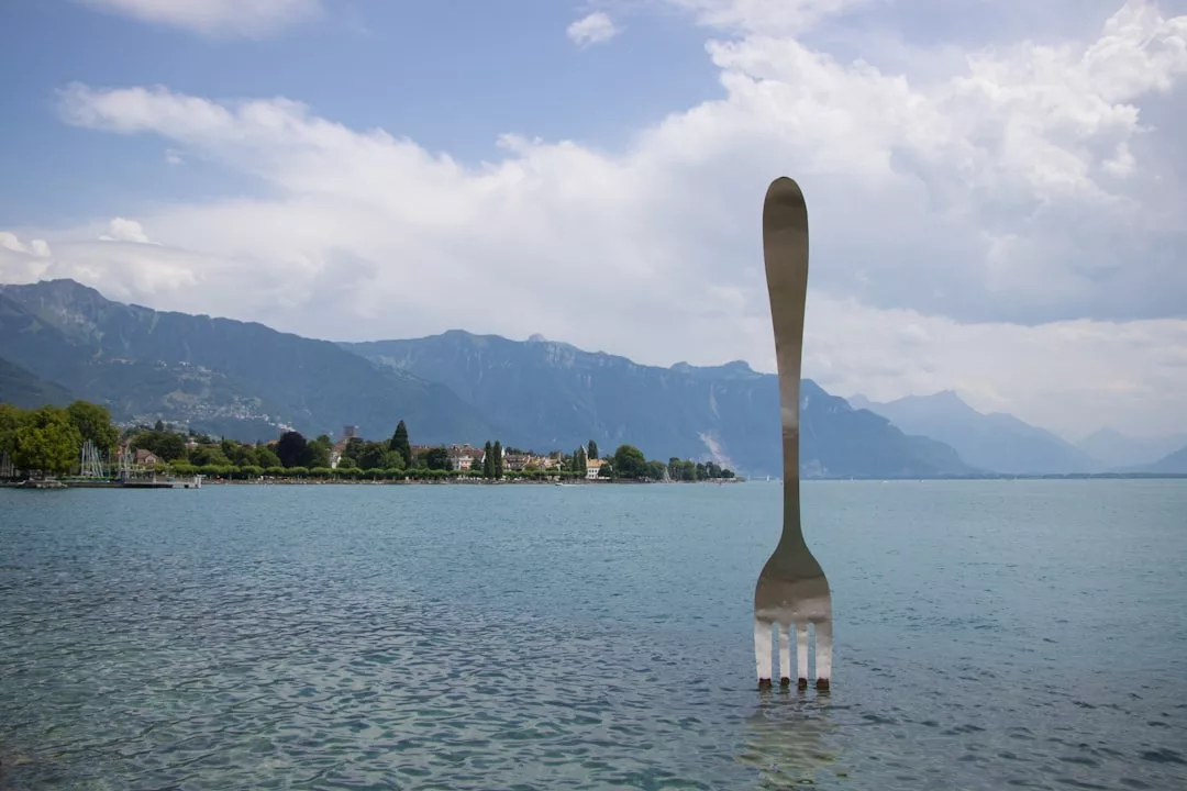 Eine große Metallgabel-Skulptur steht aufrecht in einem See, der an Kunstwerke auf Mallorca erinnert, mit Bergen und Gebäuden im Hintergrund unter einem teilweise bewölkten Himmel.